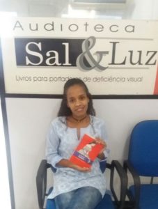 Foto de Adriana Abreu segurando o livro ao qual foi leitora sentada em uma cadeira
